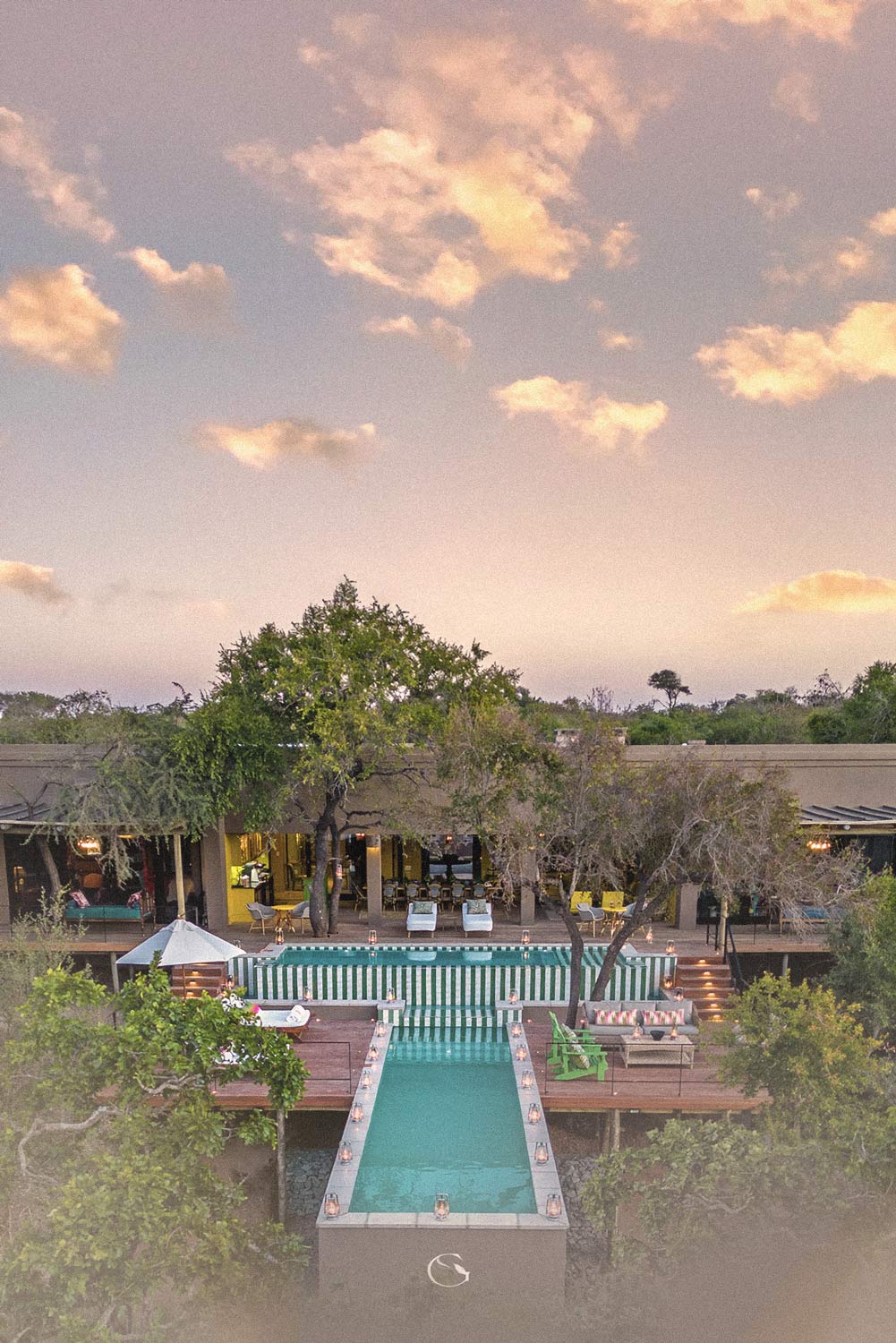 A lodge’s long pool and deck among the trees at dusk, seen from above